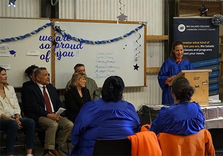 A woman in a blue graduation gown speaks at a wooden podium during a commencement ceremony held inside the Arizona State Prison Complex-Perryville. Seated in front of her are fellow graduates in identical gowns, with orange uniforms visible underneath. Behind her stands a banner for Rio Salado College, and a whiteboard decorated with blue streamers, stars, and the handwritten message “We are so proud of you!!” listing several graduate names. A group of dignitaries and supporters sit off to the side, smiling