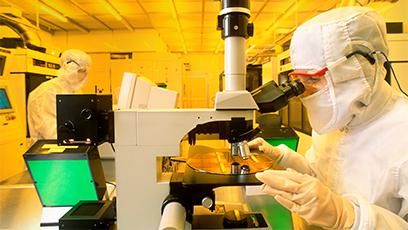 Technician using microscope to inspect silicon wafer with computer chips in production cleanroom.