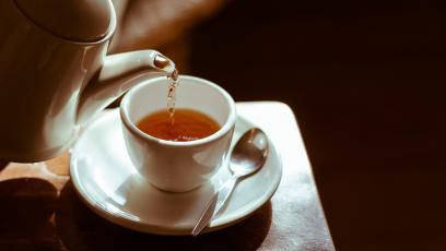 A teapot pouring tea into a cup on a saucer with a spoon on a table