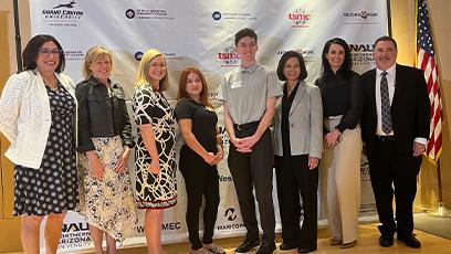 Group of eight people standing and smiling in front of a step-and-repeat banner featuring multiple company logos, including TSMC and Arizona@Work. The group includes both men and women dressed in business or business-casual attire. An American flag is visible on the right side of the image.
