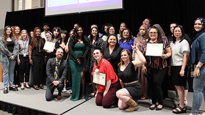 A large, diverse group of students and staff pose together on a stage at a Rio Salado College event, celebrating achievements. Several individuals proudly hold certificates, while others smile toward the camera. A woman in a sparkling green gown stands near the center. Behind the group, a projection screen displays a photo and testimonial from a student named Karen “Kiki” along with her written message.