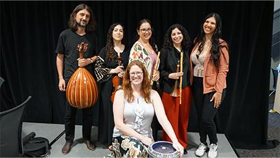 ASU’s Middle East Music Ensemble, Layali Al-Sham, pose together on a stage in front of a black curtain. Three women hold traditional Middle Eastern instruments, including a ney (flute) and a darbuka (goblet drum), while a man on the left holds an oud (lute). The seated woman in front smiles while resting her hands on the darbuka. 