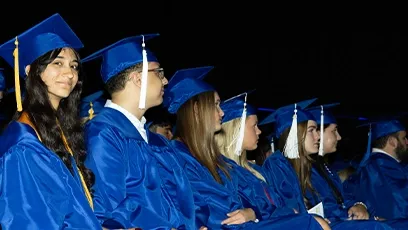 A row of graduates in royal blue caps and gowns sit side by side during a commencement ceremony. Most are looking forward, while one young woman near the front smiles at the camera. The lighting highlights the graduates against a dark background, emphasizing the celebratory moment.
