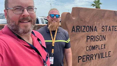 A selfie of two smiling professionals standing outdoors in front of a sandstone sign reading “Arizona State Prison Complex – Perryville.” On the left is Ken Wilk, faculty chair for applied technology and engineering, wearing glasses, a short-cropped beard, and a red polo shirt. On the right is Casey Werhanowicz, Program Director at ASPC‑Lewis, with a trimmed reddish-gold beard, sunglasses, and a dark shirt with a yellow‑striped lanyard. A palm tree and blue‑sky background appear behind them.