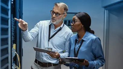 A man and a woman in professional attire stand in a server room, examining equipment. Both wear ID badges and hold tablets, collaborating while the man points at one of the servers. The environment is lit with a cool blue tone, emphasizing a high-tech workspace.