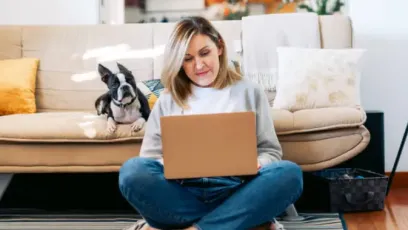 Woman at home sitting on the floor with her back against a sofa working on a laptop while her French bulldog sits by her watching her