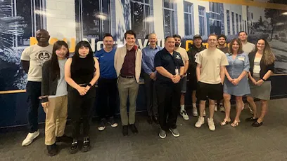 A group photo of about fifteen people standing indoors in front of a large mural wall. The group includes both men and women, casually and professionally dressed, smiling toward the camera.