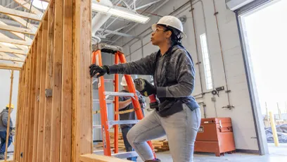 A woman wearing a white hard hat and safety gloves works on a wooden wall frame inside a construction training facility. She is using tools while standing on a platform, with ladders and other workers visible in the background.