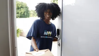 Smiling Meals on Wheels volunteer wearing a navy T-shirt delivers a boxed meal while standing at an open front door.