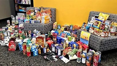 Large assortment of donated food items, including canned goods, boxed meals, snacks, and toiletries, displayed on the floor and chairs inside a building with a yellow wall.