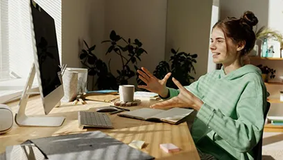 A young woman in a green hoodie sits at a desk in a sunlit home office, speaking and gesturing with her hands during a video call on a desktop computer. A notebook, keyboard, coffee mug, and tablet are arranged on the wooden desk, with houseplants and a mirror in the background.
