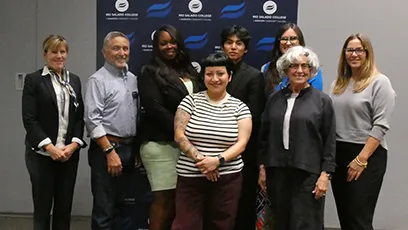 A group of eight adults stand together smiling in front of a branded backdrop, posing for a photo at a recognition event.