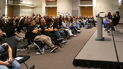 A large audience seated in a conference room facing a stage where speakers are presenting during an event.