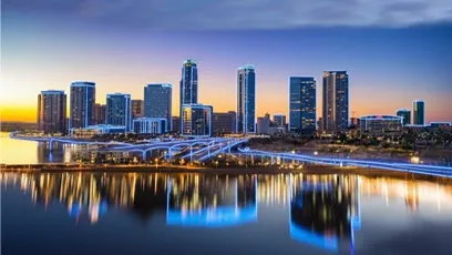 Downtown skyline at dusk with illuminated high-rise buildings and their reflections shimmering in the water along a riverfront.