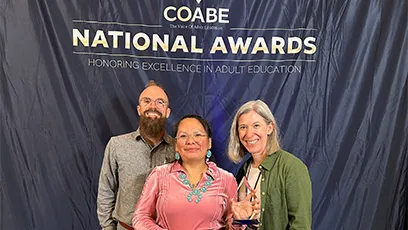 Three people stand smiling in front of a backdrop reading “COABE National Awards Honoring Excellence in Adult Education,” with one person holding an award.