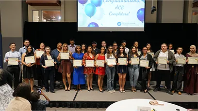 Large group of ACE program completers and staff pose on stage holding certificates, smiling in front of a screen that reads “Congratulations ACE Completers.”