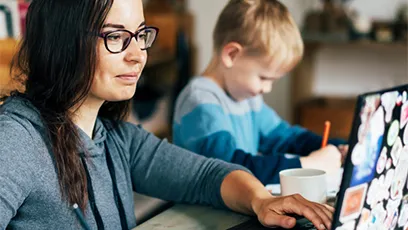 Adult learner working on a laptop at home while helping a child with schoolwork, illustrating flexible online learning in a real-life setting.