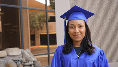 Portrait of Ebony Livas wearing a blue graduation cap and gown, standing outdoors and looking at the camera.