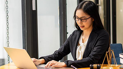 Woman wearing glasses and business attire works on a laptop at a desk, with a gavel and scales of justice nearby.