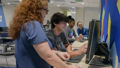 An instructor stands beside a student as they work together at a desktop computer in a classroom, while three other students use computers in the background, illustrating collaborative, technology-enabled learning.