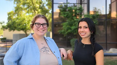 Maren “Rain” Colon, left, and Ariel Davila, right, smile together outdoors in front of a glass building and greenery.