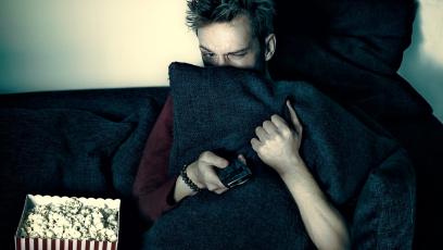 Scared teen hiding behind pillow sits on couch with bucket of popcorn, holding a remote
