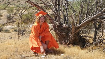 photo of Emma Harlow in a desert landscape kneeling under a dead tree in her graduation regalia.