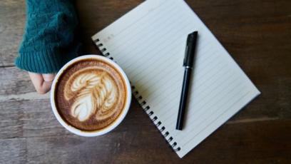 A hand holding a latte next to a notebook with a pen