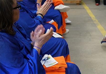 A group of individuals in blue graduation gowns and orange pants are seated in a row, clapping. The focus is on their hands and laps, with one person holding a colorful program booklet and folded orange fabric. The scene is part of a graduation ceremony taking place at Arizona State Prison Complex-Perryville