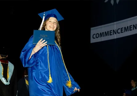 Madelyn Bailey, a senior at Desert Vista High School and dual enrollment student, smiles proudly as she walks across the stage in her blue graduation cap and gown. She holds up her Rio Salado College diploma cover while wearing an honor cord, with a large "Commencement" banner hanging in the background and faculty members visible off to the side.