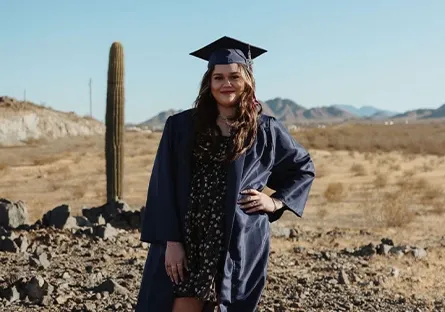 YAz J., a graduate from Heritage Academy Maricopa, stands confidently in a desert landscape wearing a dark graduation gown and cap. With one hand on her hip, she smiles at the camera against a backdrop of rocky terrain, a tall saguaro cactus, and distant mountains.