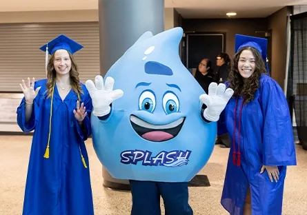 Madelyn Bailey and Alexandria Woodruff, dual enrollment students in blue graduation gowns and caps, pose on either side of Splash, the Rio Salado College mascot. All three are smiling and waving at the camera indoors before the commencement ceremony. Splash, a cheerful blue water droplet character, stands in the center with raised white-gloved hands, adding a fun and festive touch to the occasion.