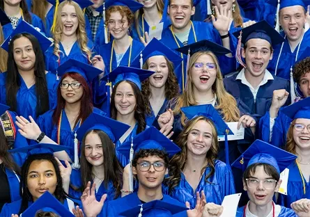 A large, energetic group of dual enrollment students from Rio Salado College cheer and smile during commencement. They are dressed in bright blue graduation gowns and caps, some holding diplomas and raising their hands in celebration. The diverse group of young graduates stands close together, radiating excitement and pride as they mark this academic milestone.
