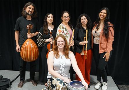 ASU’s Middle East Music Ensemble, Layali Al-Sham, pose together on a stage in front of a black curtain. Three women hold traditional Middle Eastern instruments, including a ney (flute) and a darbuka (goblet drum), while a man on the left holds an oud (lute). The seated woman in front smiles while resting her hands on the darbuka.