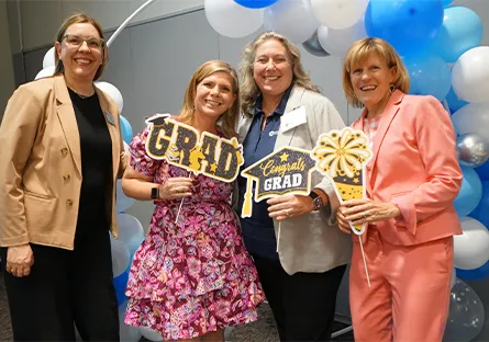 Four women smile and pose together at a graduation celebration, holding festive signs that say “GRAD” and “Congrats GRAD.” They stand in front of a balloon arch with blue, white, and silver balloons. The atmosphere is joyful and celebratory, with one woman wearing a bright pink floral dress and others dressed in business casual attire.