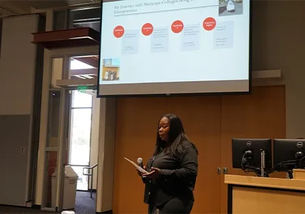 Shaniqua Hopkins, founder of the nonprofit 7th Dream Helping Hearts, presents at the front of a conference room with a microphone and printed notes in hand. Behind her, a large screen displays a slide titled “My Journey with Maricopa Colleges to Being an Entrepreneur.” The room is softly lit, and the speaker stands near a wooden podium with dual computer monitors.