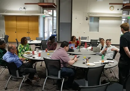 A group of people sit around two round tables in a conference room at Rio Salado College, engaged in discussion during a small business networking event. Soft drinks and snacks are on the tables, and one participant speaks while others listen attentively. The atmosphere is casual and collaborative.