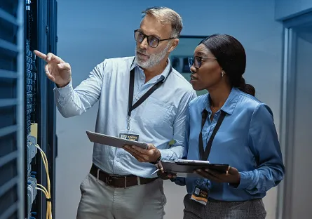 A man and a woman in professional attire stand in a server room, examining equipment. Both wear ID badges and hold tablets, collaborating while the man points at one of the servers. The environment is lit with a cool blue tone, emphasizing a high-tech workspace.