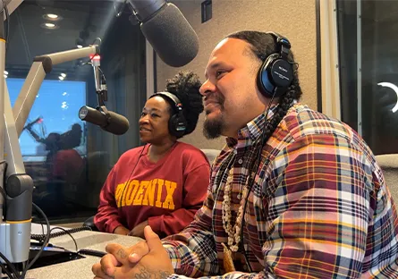 Two guests sit in a radio studio during the recording of the Wave Makers Podcast. Wearing headphones and seated in front of large microphones, they appear engaged in conversation. The man in the foreground wears a plaid shirt and beaded necklace, while the woman behind him wears a red “Phoenix” sweatshirt and has her hair styled in a high puff. The studio setting features soundproof walls and recording equipment.