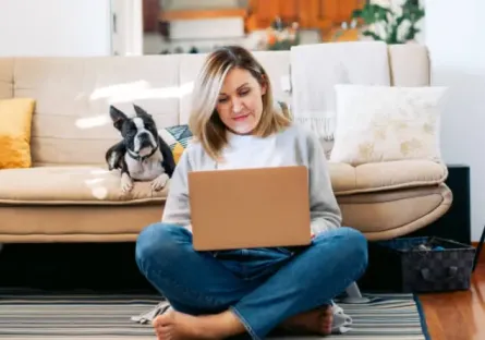 Woman at home sitting on the floor with her back against a sofa working on a laptop while her French bulldog sits by her watching her