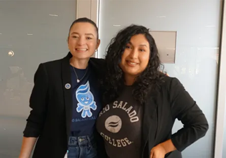 Two Rio Salado College employees smile and pose together indoors. Both are wearing black blazers; one has on a Rio Salado College T-shirt, while the other wears a shirt featuring the Splash mascot. They stand in front of a frosted glass wall, looking friendly and approachable.
