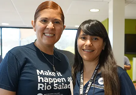 Two Rio Salado College employees smile together inside a brightly lit office space. The employee on the left wears a navy T-shirt with the words “Make it happen at Rio Salado” printed on the front, while the employee on the right wears a Rio Salado College polo shirt and a lanyard decorated with the phrase “Star Wars.” Sunlight shines through large windows in the background, giving the image a warm and welcoming feel.