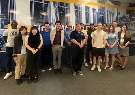 A group photo of about fifteen people standing indoors in front of a large mural wall. The group includes both men and women, casually and professionally dressed, smiling toward the camera.