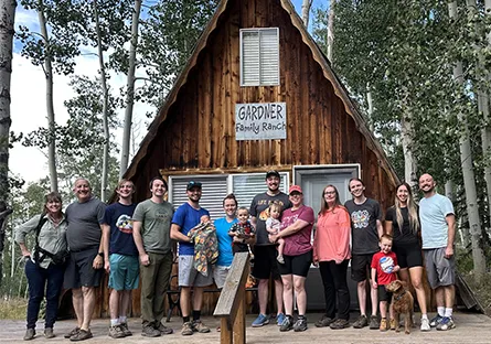 A family group stands together on the wooden porch of a rustic A-frame cabin with a sign reading “Gardner Family Ranch.” The group includes adults, young children, and a dog, all smiling at the camera. The cabin is surrounded by tall aspen trees.
