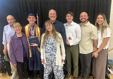 A young graduate wearing a blue cap and gown with multiple honor cords, medals, and a diploma smiles while standing with his family. Eight family members of different generations gather closely around him, smiling proudly for the photo. They are posed indoors in front of a black curtain, celebrating his academic achievement together.