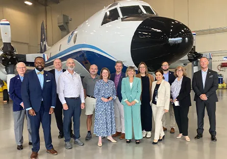 A group of thirteen people stand together inside an aircraft hangar, smiling at the camera. Behind them is a large white and blue propeller airplane. The group includes men and women dressed in business and business-casual attire.