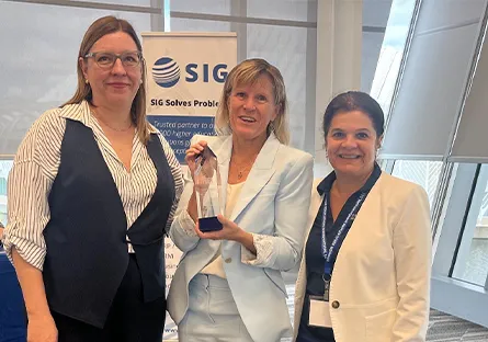 Three women stand together indoors, smiling for a photo at an event. The woman in the center, wearing a light blue suit, holds a glass award. She is flanked by two colleagues—one on the left in a striped blouse with a dark vest and trousers, and one on the right in a white blazer and blue pants. A banner behind them displays the SIG logo and tagline “SIG Solves Problems,” indicating the award recognition took place at a professional conference or gathering.