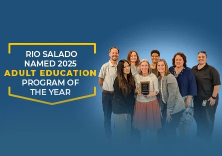 A group of Rio Salado College staff members stand together smiling, with one person in the center holding a plaque. The background features a blue gradient with white and yellow text that reads: “Rio Salado Named 2025 Adult Education Program of the Year.” The team poses proudly, representing the college’s award-winning adult education program.
