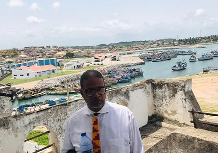 A man wearing glasses, a white shirt, and a colorful patterned tie stands outdoors holding a water bottle. Behind him is a historic white stone structure overlooking a harbor filled with fishing boats and buildings under a bright, partly cloudy sky.