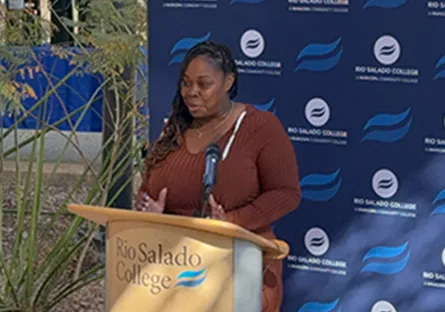 Shaniqua Hopkins speaks at a podium with the Rio Salado College logo, set against a blue backdrop with the college’s branding. A banner beside her reads “Empower[ED] for Student Success,” highlighting support for student opportunities. The outdoor event setting includes plants and natural light, emphasizing community engagement and educational empowerment.